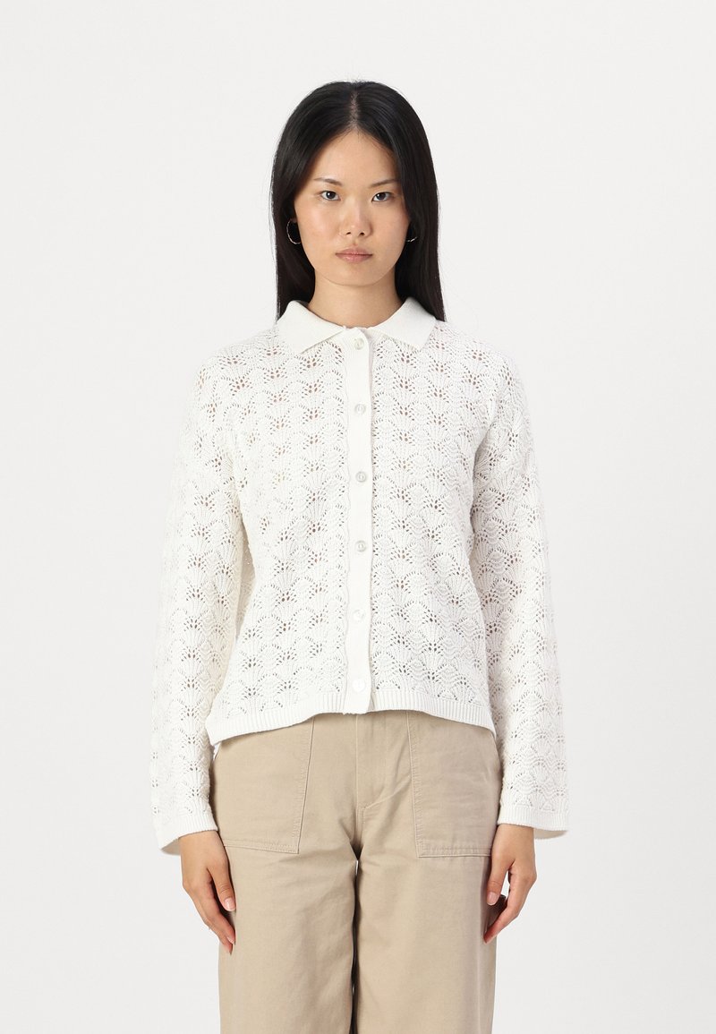 Bela pletena kardigan s čipkastim vzorcem, z gumbi spredaj, rebrastim ovratnikom in manšetami. V kombinaciji z bež hlačami, ki prikazujejo sproščenega obliko.
