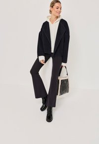 Veste noire cropped avec un large col, associée à un hoodie blanc, un pantalon noir ajusté et des bottes noires brillantes ; sac noir avec des accents en peluche.