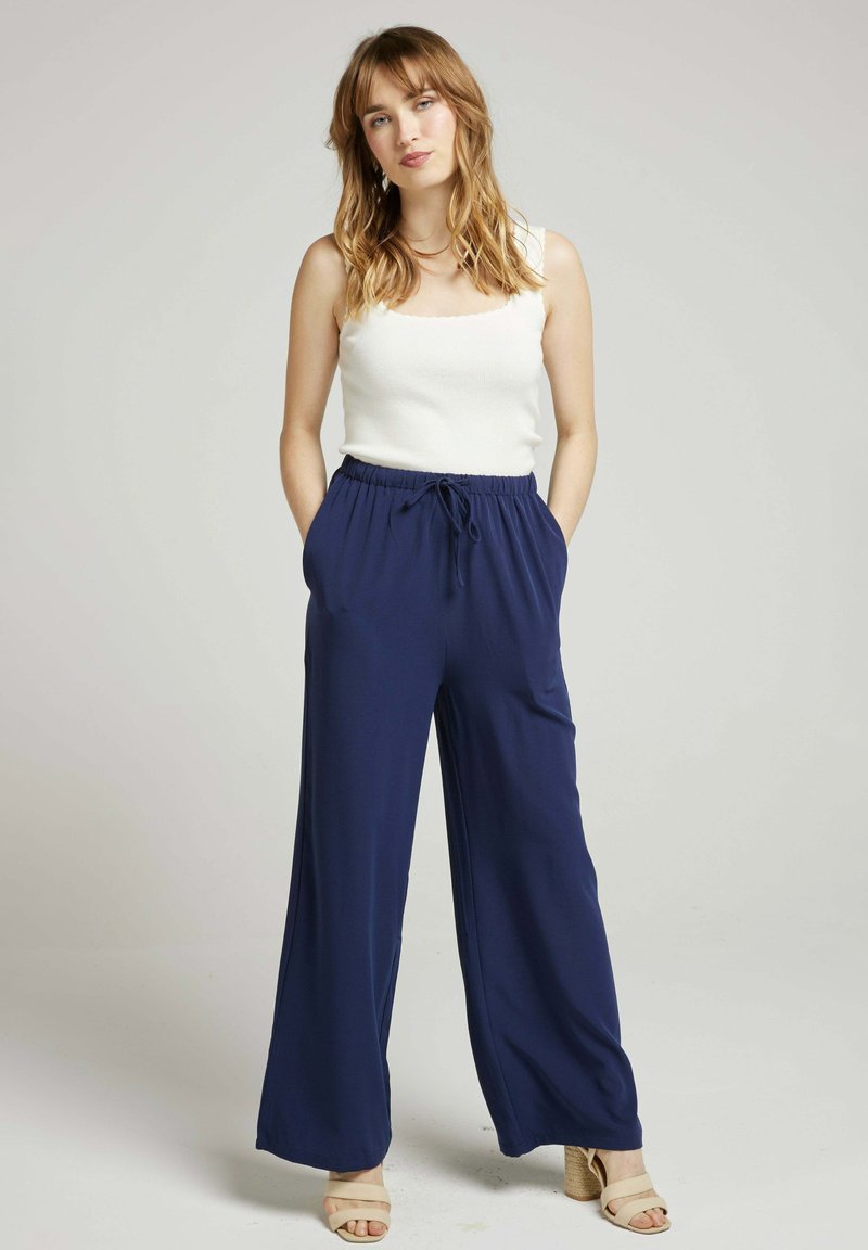 Pantalons larges bleu marine en tissu léger avec une taille élastique et un cordon de serrage. Associés à un top sans manches blanc et des sandales neutres.