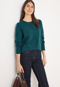 Maglione in maglia teal con scollatura rotonda e polsini a costine, abbinato a jeans di denim scuri; tiene in mano una borsa marrone con dettagli in metallo.