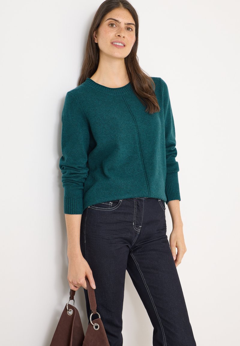 Maglione in maglia teal con scollatura rotonda e polsini a costine, abbinato a jeans di denim scuri; tiene in mano una borsa marrone con dettagli in metallo.