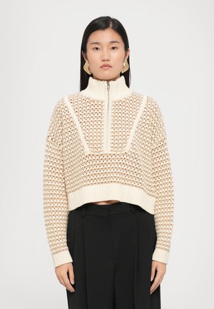 Maglione accorciato color crema con collo alto, chiusura a metà lampo e motivo a maglia aperta texturizzato. Abbinato a pantaloni neri.