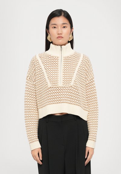 Maglione accorciato color crema con collo alto, chiusura a metà lampo e motivo a maglia aperta texturizzato. Abbinato a pantaloni neri.