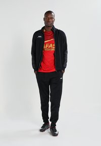 Giacca nera con zip e logo, maglietta sportiva rossa con scritte gialle, pantaloni neri e scarpe da calcio nere con accenti bianchi e dettagli rossi.