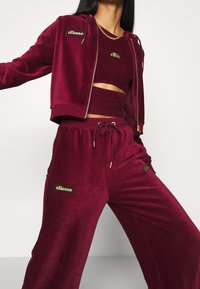 Modelo que lleva un chándal de terciopelo burdeos con chaqueta con cremallera, top corto y pantalones de pierna ancha que presentan logotipos de ellesse en dorado y negro.