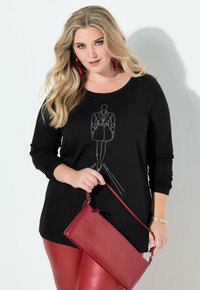 Blondýna s vlnitými vlasy má na sobě černý top s motivem kabátu ozdobeným kamínky, červené kožené legíny, drží červenou kabelku a nosí červené náušnice.