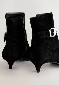 Bottines en suède noir avec un talon effilé, dotées d'une fermeture éclair sur le côté et d'une sangle décorative à boucle autour de la cheville. Texture lisse.