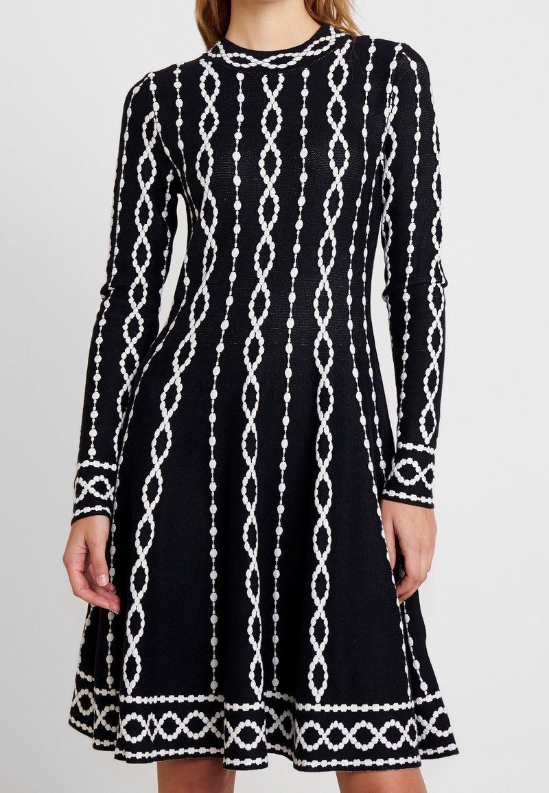 Robe en maille noire à manches longues, ornée de motifs décoratifs blancs composés de formes perlées alternées et de lignes, évasée à l'ourlet.