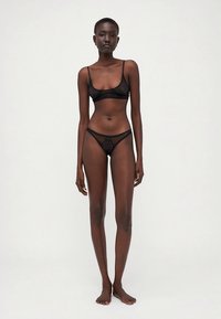 Conjunto de sutiã bralette e tanga em renda preta com tecido transparente, bordas em cascata e detalhes bordados. Design simples com poucos elementos metálicos.