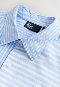 Chemise boutonnée à rayures bleu clair et blanches avec un col et une étiquette noire portant l'inscription « NEXT EDIT ». 