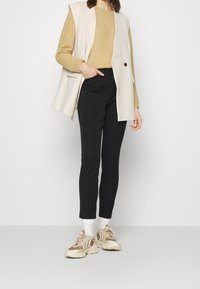 Veste sans manches beige superposée sur un pull moutarde, associée à un pantalon noir et des baskets marron clair avec des semelles et lacets blancs.
