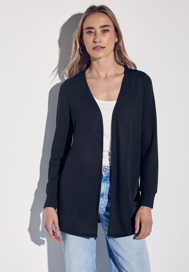 Lätt, svart cardigan utan knäppning med långa ärmar och ribbade manschetter, lagd över en vit topp, tillsammans med ljusblå jeans.