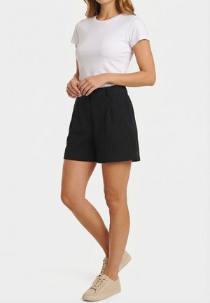 Femme portant un haut blanc à manches courtes, un short noir taille haute et des baskets beige sur un fond uni.