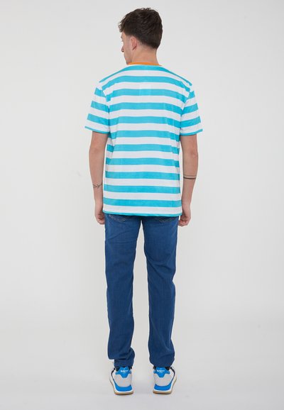 T-shirt a righe nei toni del blu chiaro e bianco, maniche corte, con un accento arancione sullo scollo, abbinata a jeans blu e sneakers multicolori.