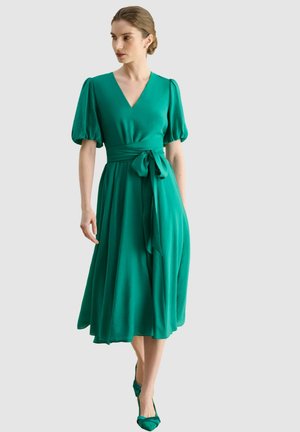 Femme portant une robe midi verte avec des manches bouffantes et une ceinture nouée à la taille, associée à des talons verts assortis, debout devant un fond uni.