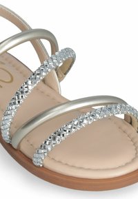 Sandalia con tiras que presenta bandas metálicas plateadas adornadas con strass transparentes, una plantilla acolchada de color beige y una suela marrón texturizada.