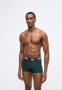 Μυώδης άνδρας όρθιος, φοράει σκουροπράσινα μπόξερ Lacoste με μαύρη μέση και λευκό λογότυπο σε απλό φωτεινό φόντο.