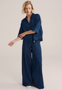 Džínová oversized košile s límečkem, knoflíkovým zapínáním a ohrnutými rukávy, doplněná širokými džínovými kalhotami tmavě modré barvy.