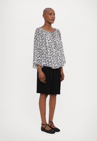 Lett, gjennomsiktig bluse med blomstermønster i svart og hvitt, kombinert med svart shorts til kneet og svarte flate sko med prikkete design.