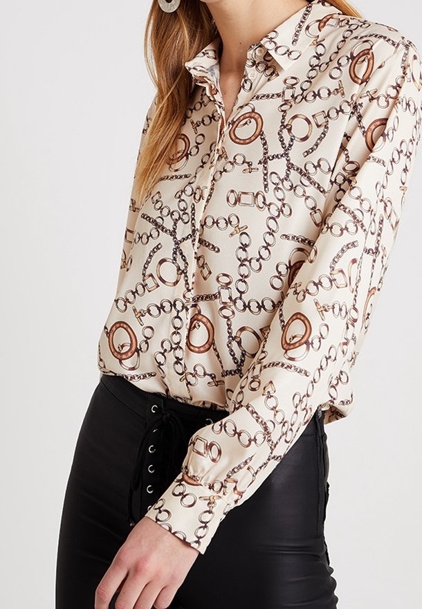 Blouse couleur crème avec un motif en maillons de chaîne brun et doré. Présente un col, une fermeture à boutons et de longues manches.