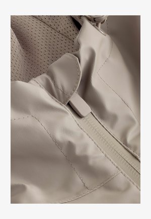 Svijetlo bež jakna s teksturiranom, prozračnom unutrašnjom podlogom. Ima glatku vanjštinu, sakupljenu ovratnik i diskretan zatvarač na munje.