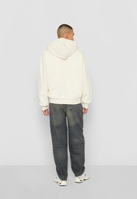 Hoodie crème avec une grande capuche structurée, coupe décontractée et poignets à côtes, associé à un jean bleu foncé ample et des baskets blanches.