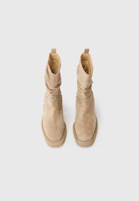 Bottes mi-mollet en suédine beige avec une tige fléchie, bout rond, semelle épaisse et petits strass décoratifs autour de la cheville.