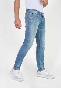 Jeans de mezclilla de un azul claro con corte ajustado, que presentan un diseño clásico de cinco bolsillos, detalles sutiles de desgaste y una etiqueta de cuero marrón en la cintura.