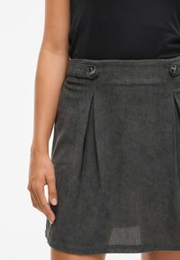 Vrouw in een donkergrijze corduroy rok met knoopsluitingen en verticale plooien, gecombineerd met een zwarte mouwloze top.