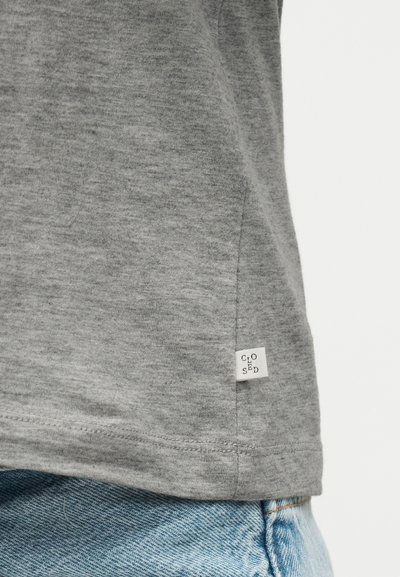 Πολύχρωμο γκρι τελείωμα T-shirt με μικρή λευκή ετικέτα που γράφει "CLOSED" με μαύρα γράμματα πάνω από ανοικτό μπλε τζιν.