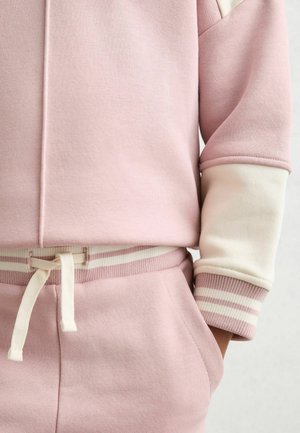 Nærbillede af pink og cremefarvet sporty outfit med snøre i taljen, stribet manchet og hånd i lommen.