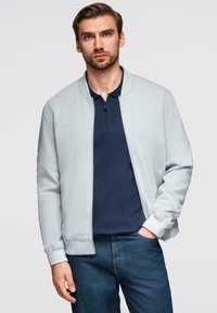 Giovane uomo con giacca grigia chiara a zip sopra una maglietta blu navy, che indossa jeans blu, in piedi con una mano in tasca contro uno sfondo semplice.