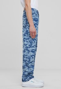 Blue camo-patterned jeans made of denim, featuring a straight leg design, five pockets, and a mid-rise waist, paired with white sneakers.