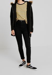 Sort parka med faux fur hætte, gul og hvid stribet skjorte, sorte skinny jeans og sorte sneakers med hvide såler.