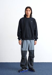 Um jovem está encostado a uma parede simples, vestindo um sweatshirt preto da Nike, calças cinza e pretas, e sapatilhas pretas, com uma expressão neutra.