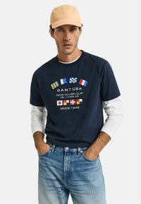 Tume puuvillane T-särk valgete pikade varrukatega all, millel on esiküljel erksad mereteemalised lipud ja tekst "GANT USA". Seljas helesinised teksad.