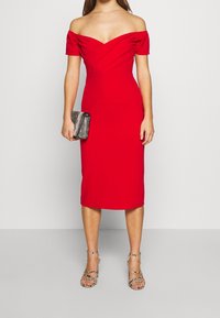 Robe rouge décolletée d'une épaule avec une silhouette ajustée, une longueur au genou et un détail de décolleté torsadé. Association avec une pochette argentée texturée et des talons à lanières.