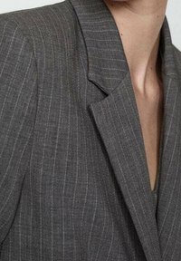 Blazer grigio con righe, collo a revers, vestibilità su misura e texture liscia, che mostra una leggera lucentezza e un design strutturato.
