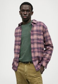 Koszula flanelowa w kratę w odcieniach fioletu i różu, nałożona na zieloną koszulkę; zestawiona z khaki spodniami z łatami i kieszeniami z przodu.