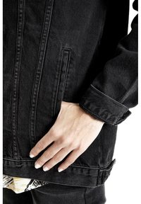 Czarna dżinsowa kurtka z widoczną kieszenią, pokazana z ręką opartą na dolnym wykończeniu. Tkanina ma teksturowane wykończenie i nieco wyblakły wygląd.