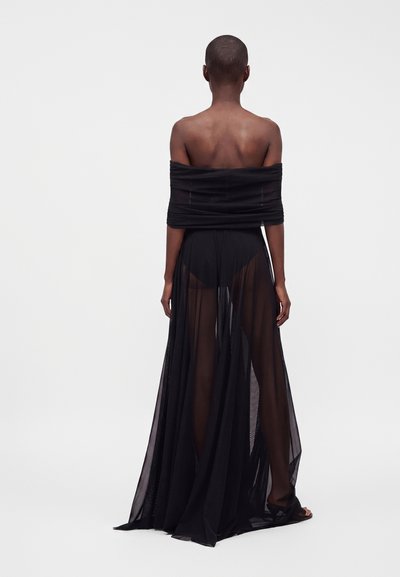 Femme aux cheveux courts portant une robe noire transparente épaules dénudées, debout pieds nus, dos à la caméra sur un fond uni.