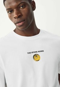 Valkoinen puuvillapaita, jossa on pyöreä keltainen graafinen kuva mustalla tekstillä "THE STONE ROSES" rinnassa.