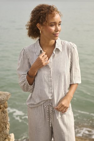 Naine lokkis juustega, kandes triibulist särki ja pükse, seisab kivi ja rahuliku mere lähedal, vaadates küljele õrna naeratusega.