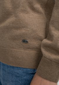 Brauner Strickpullover mit gerippten Bündchen und Saum, versehen mit einem kleinen silbernen Etikett mit der Aufschrift "Petrol". Über blauer Denim-Stoff getragen.