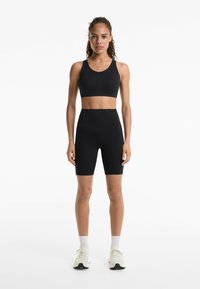 Čierny športový crop top a cyklistické šortky s vysokým pásom v rebrovanej textúre. Modelka má na sebe svetlé topánky a členkové ponožky, s neutrálnym pozadím.