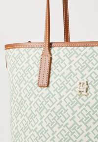 Tote bag med et grønt geometrisk mønster på en kremfarget bakgrunn, brune lærdetaljer og en gulllogo. Glatt tekstur og strukturert fasong.