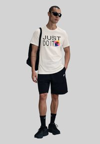 Camiseta color crema con el texto "JUST DO IT" y el logo de Nike, combinada con pantalones cortos negros y zapatillas negras. Los accesorios incluyen gafas de sol y una mochila.