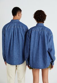 Jeansskjorte i medium blå, med klassisk krage, knappeender og en løs passform. Båret av to personer fra bak.