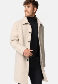 Beige uldblandingsfrakke med lige skæring, spidse krave og sorte knaplukninger. Bar den over en sort turtleneck og sorte bukser.
