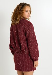 Suéter burdeos texturizado con acentos brillantes, puños y dobladillo acanalados, combinado con una falda ajustada a juego, que muestra un ajuste relajado y un diseño tejido.